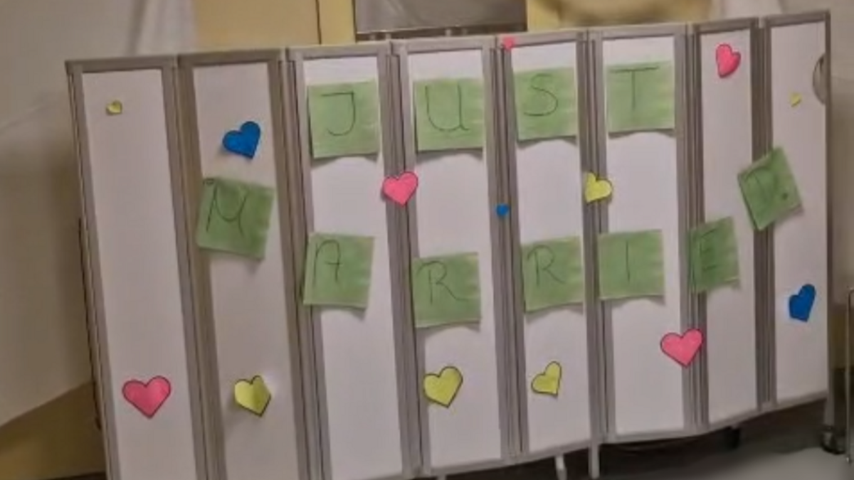 Auf einer Stellwand sind grüne Zettel mit Buchstaben, die das Wort Just Married ergeben, rundherum sind Herzen dekoriert. Im Hintergrund sieht man eine Girlande aus Ringen.